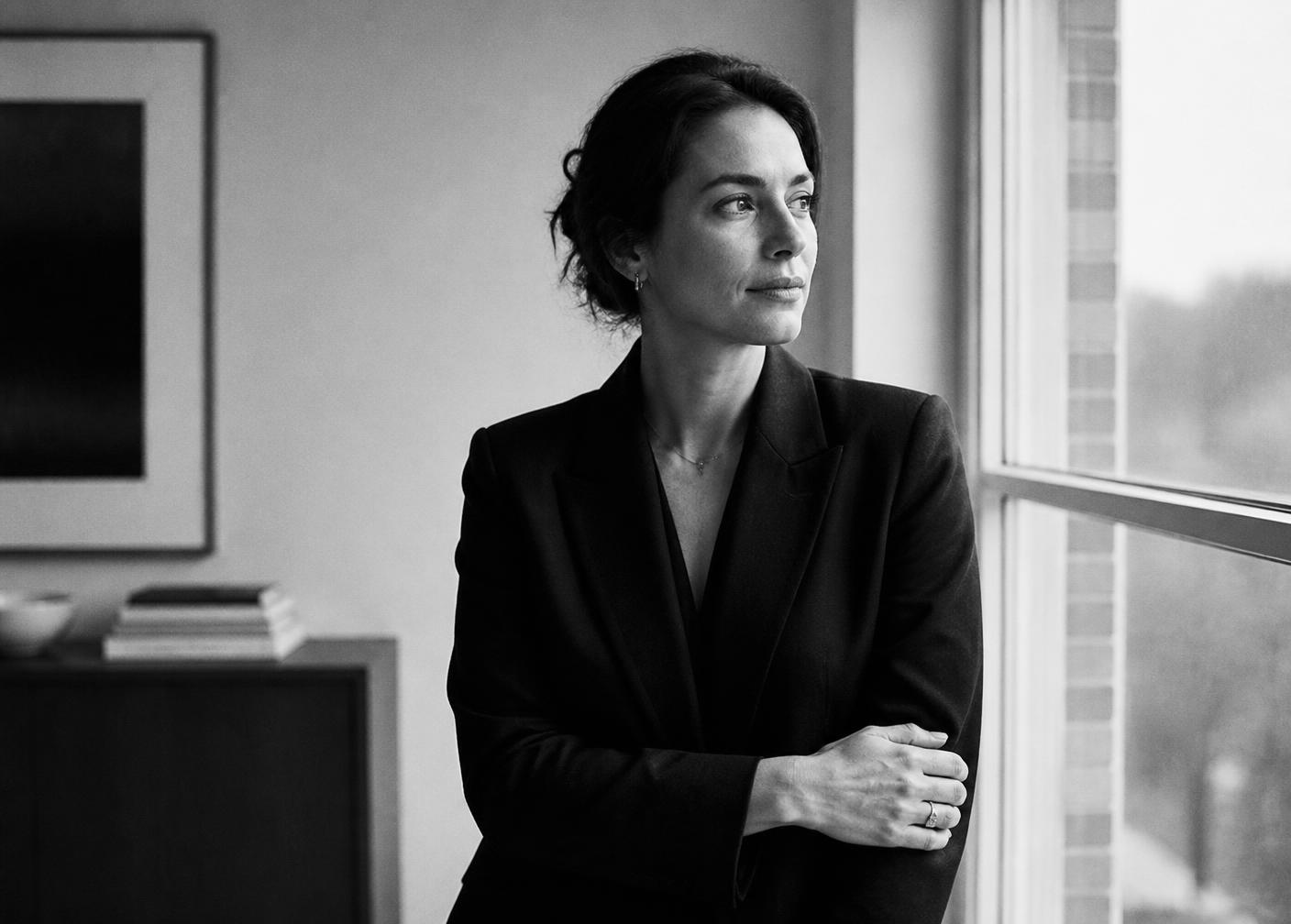 A woman in a blazer standing by a window in quiet daylight